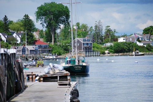 A calm harbor scene with a sailboat named "Tetra" docked alongside wooden piers and quaint houses in the background.