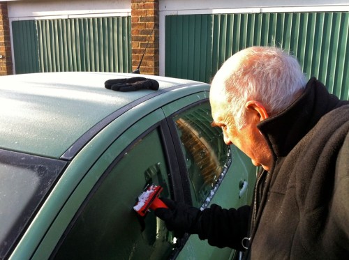 An older man scrapes frost off a green car window on a cold morning, wearing a black coat and glove.