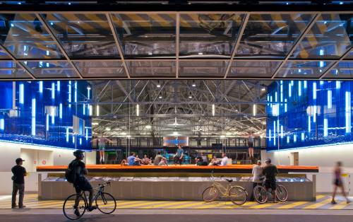 A modern building with a glass facade, featuring people inside and bicycles parked outside, illuminated by blue lights.