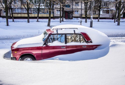 A red car partially buried in deep snow, surrounded by snow-covered trees and buildings in a winter scene.