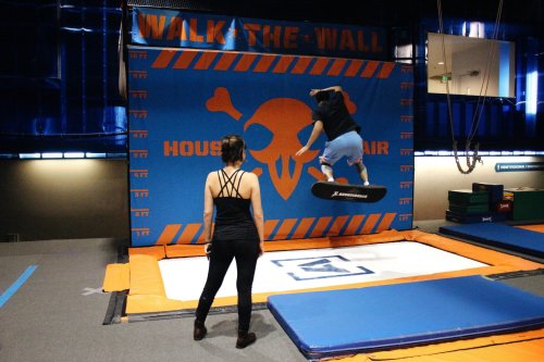 A person jumps on a trampoline towards a wall with a skull logo, while another person watches nearby.