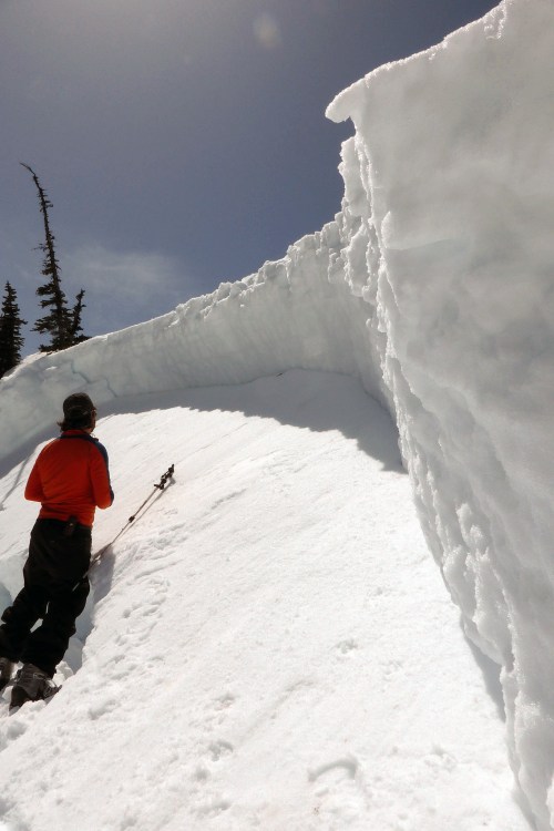 A person in an orange jacket stands on a snowy slope, looking up at a tall snowbank under a clear blue sky.