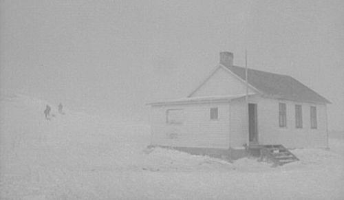A small white house in a snowy landscape, partially obscured by fog, with two figures in the distance.