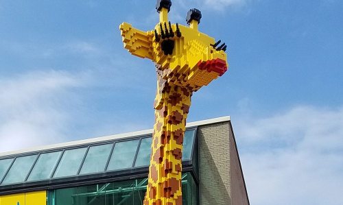 A large giraffe sculpture made of colorful LEGO bricks stands outside a building under a blue sky.
