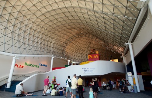 Interior of a children's museum with a high, geometric ceiling and colorful signage promoting play, discovery, and learning.