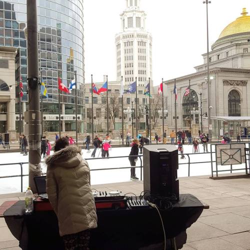A DJ plays music near an outdoor ice skating rink, with flags and buildings in the background.
