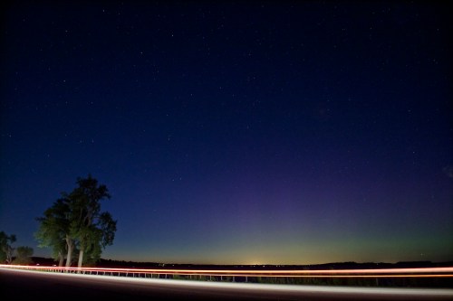 A serene night sky filled with stars, featuring a faint aurora and light trails from passing vehicles along a road.