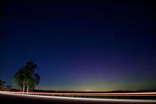 A serene night sky filled with stars, featuring a faint aurora and light trails from passing vehicles along a road.