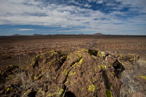 A rocky outcrop covered in moss overlooks a vast, arid landscape with distant mountains under a cloudy sky.
