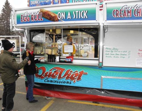A food concession stand with a sign for "Giant Egg Roll on a Stick" and a customer ordering from the vendor.