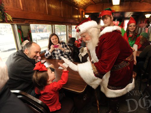 A cheerful Santa interacts with a young girl at a festive gathering, surrounded by smiling families and elves.