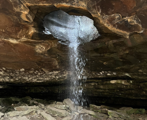 Water cascades from a hole in a rocky cave ceiling, creating a small waterfall onto the stone floor below.