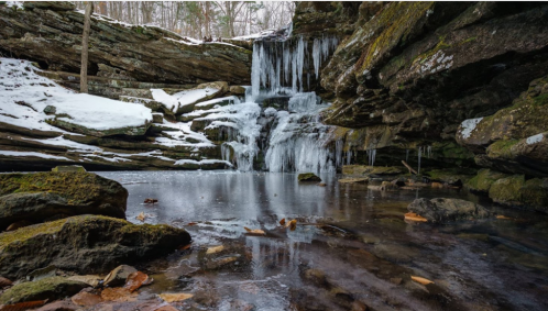 A serene winter scene featuring a frozen waterfall cascading over rocks into a still pool, surrounded by snow and trees.
