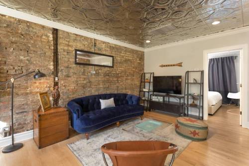 Cozy living room with a blue velvet sofa, exposed brick wall, wooden furniture, and a TV on a stand.