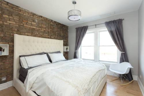 Cozy bedroom with a tufted bed, brick wall, large windows, and elegant gray curtains.