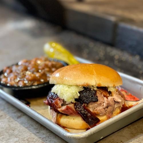 A pulled pork sandwich with coleslaw and barbecue sauce, served with a side of baked beans on a metal tray.