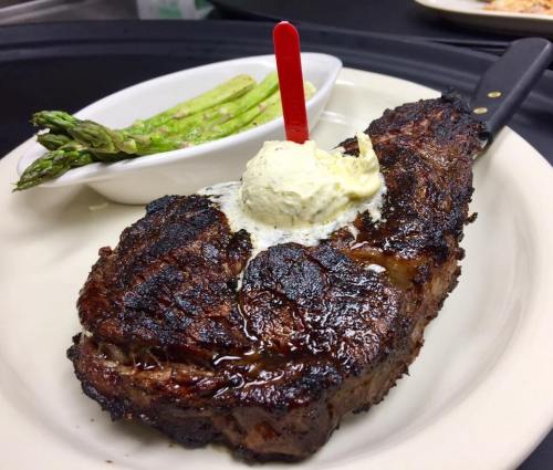 A grilled steak topped with butter and a red pick, served with a side of asparagus on a white plate.