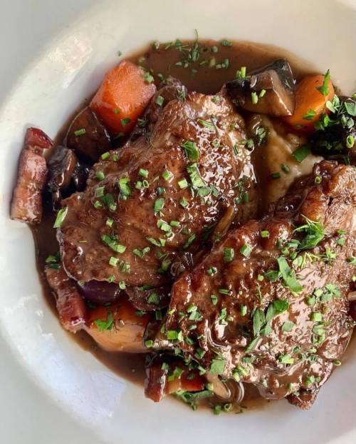 Two pieces of meat in a rich sauce, garnished with herbs, served with colorful vegetables on a white plate.