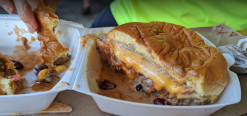 A close-up of a messy cheeseburger in a takeout container, with a hand reaching for a piece of bread.