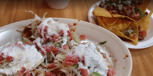 A plate of tacos topped with lettuce, tomatoes, and sour cream, alongside a serving of tortilla chips and beans.