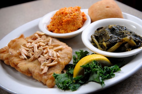 A plate featuring fried fish topped with almonds, collard greens, sweet potato mash, and a roll, garnished with lemon and kale.