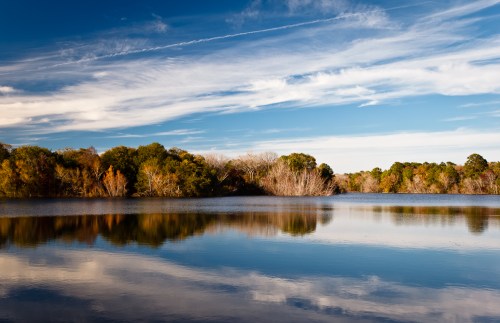 A serene lake reflecting trees and a blue sky with wispy clouds, showcasing autumn colors along the shoreline.
