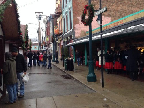A festive street scene with people shopping and enjoying holiday decorations, featuring shops and a light drizzle.