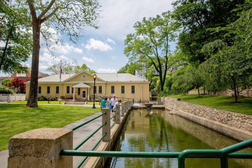 A yellow building beside a calm waterway, with people walking along a path and trees in the background under a blue sky.