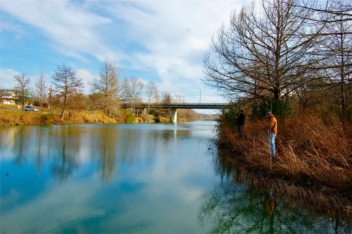 A serene river scene with a bridge in the background and a person standing by the water's edge among tall grasses.