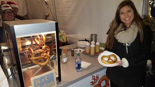 A woman holds a plate of pretzels in front of a pretzel stand, with sauces and a festive background.
