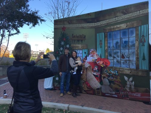 A family poses with Santa in front of a festive backdrop while another person takes a photo.