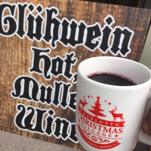 A hand holding a mug of glühwein in front of a wooden sign that reads "Glühwein hot mulled wine."