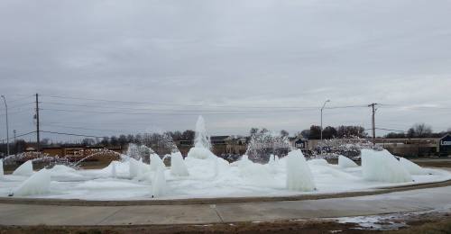 A frozen fountain with ice and water jets, set against a cloudy sky and a suburban backdrop.