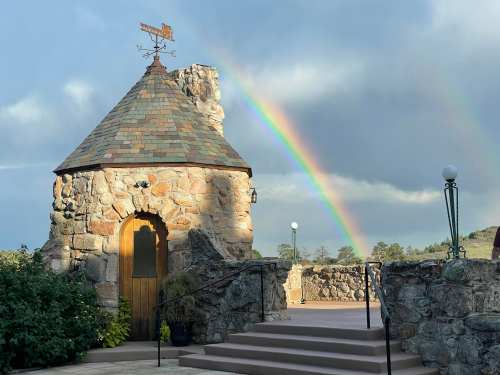 A stone building with a conical roof, surrounded by greenery, under a rainbow and cloudy sky.