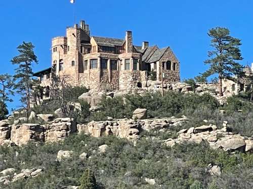 A large stone castle sits atop a rocky hill, surrounded by trees and a clear blue sky.