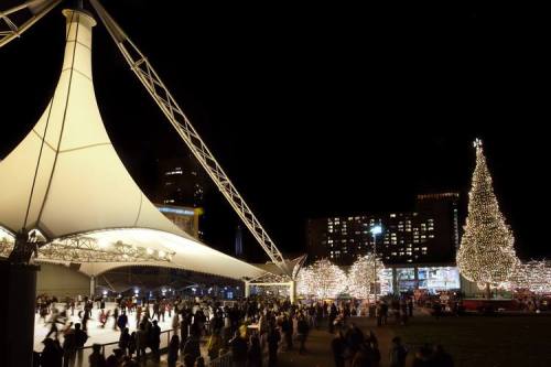 A festive outdoor scene at night with a large Christmas tree, ice skaters, and illuminated buildings.