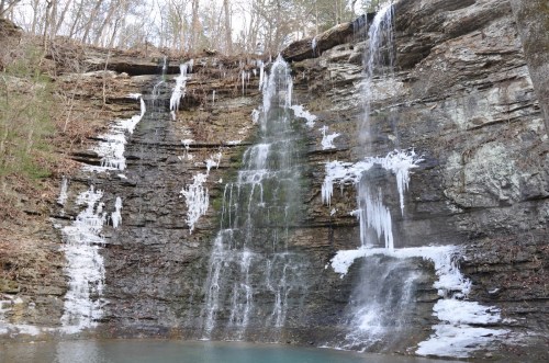 A scenic waterfall cascading down rocky cliffs, with icicles hanging and a serene pool at the base.