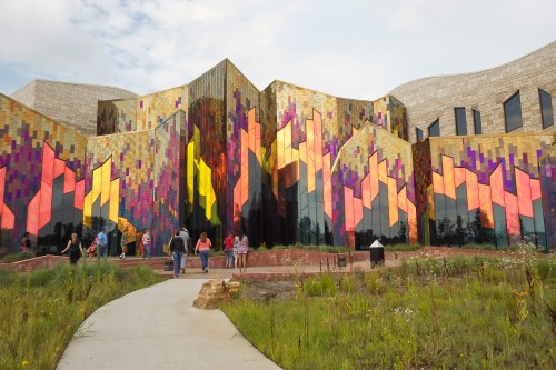 A colorful, modern building with reflective surfaces and geometric patterns, surrounded by greenery and people walking.