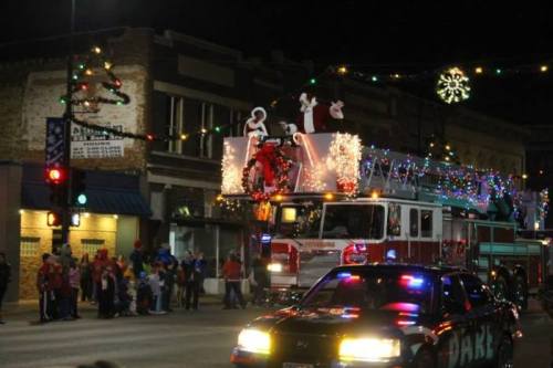 A festive fire truck adorned with lights and decorations, carrying Santa and elves during a nighttime parade.