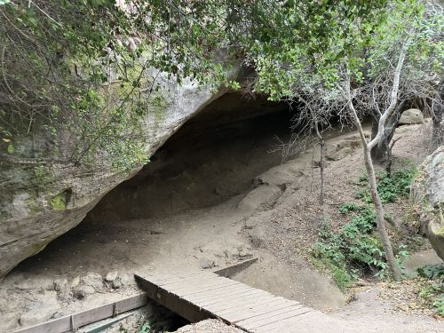 A rocky cave entrance with a wooden bridge leading into a shaded area surrounded by trees and foliage.