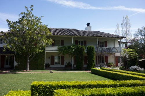 A two-story house with a tiled roof, surrounded by greenery and neatly trimmed hedges under a clear blue sky.