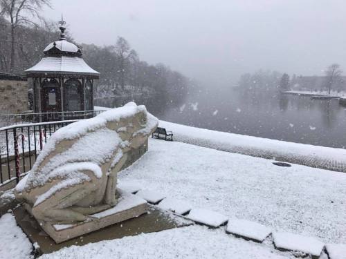 A snowy scene featuring a large frog statue by a river, with a gazebo in the background and falling snowflakes.