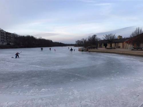 People ice skating on a frozen lake, with trees and buildings in the background under a cloudy sky.