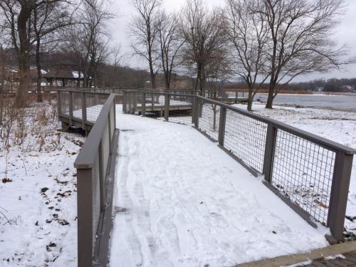 A snowy pathway leads to a lake, surrounded by bare trees and a wooden railing. Overcast sky above.