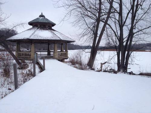 A snow-covered gazebo beside a frozen lake, surrounded by bare trees and a peaceful winter landscape.