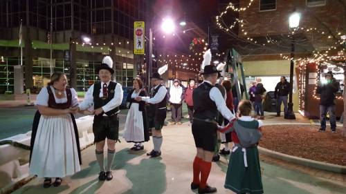 A group of people in traditional attire dancing outdoors, surrounded by festive lights and a lively crowd.