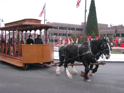 A horse-drawn trolley with passengers rides past a festive holiday scene, featuring flags and a decorated tree.
