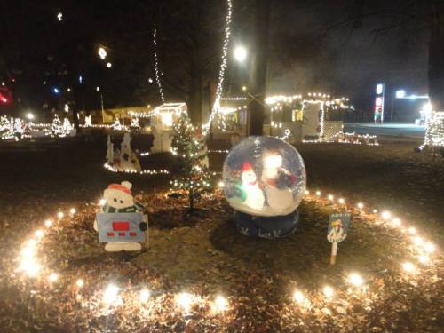 A festive scene with a snow globe, snowman, Christmas tree, and lights in a park decorated for the holidays.