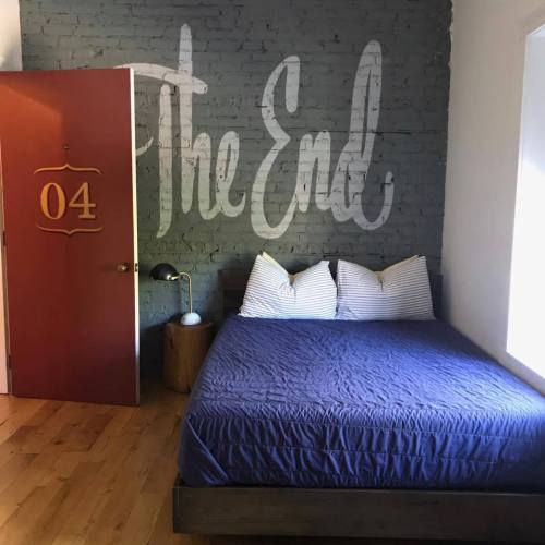 A cozy bedroom with a blue bed, striped pillows, and a door marked "04" against a gray wall with "The End" written on it.