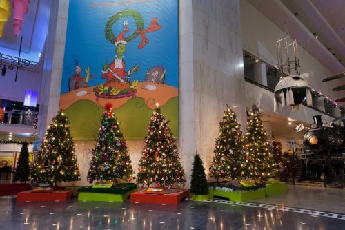 Colorful Christmas trees decorated with lights and ornaments, set against a large mural of the Grinch in a festive space.